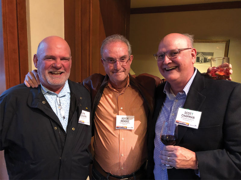 Three attendees indoors with name badges, smiling; two hold drinks.